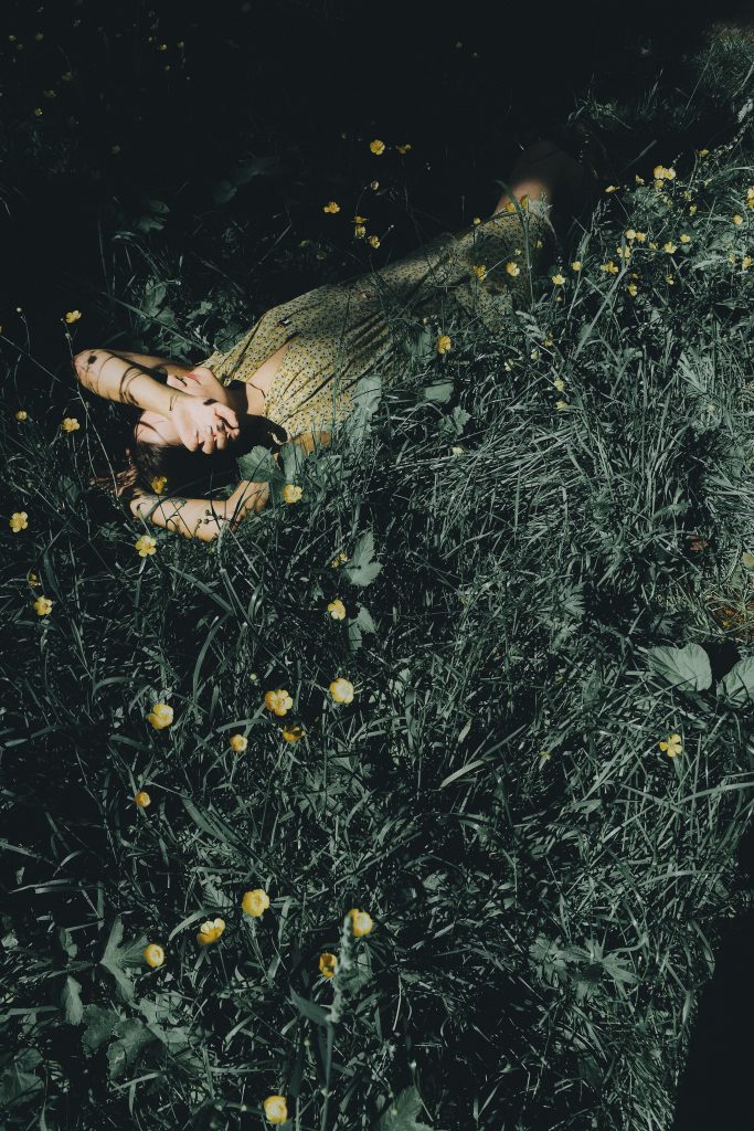 A photo of a person lying on their back in a field of tall grass with their left arm covering their face.