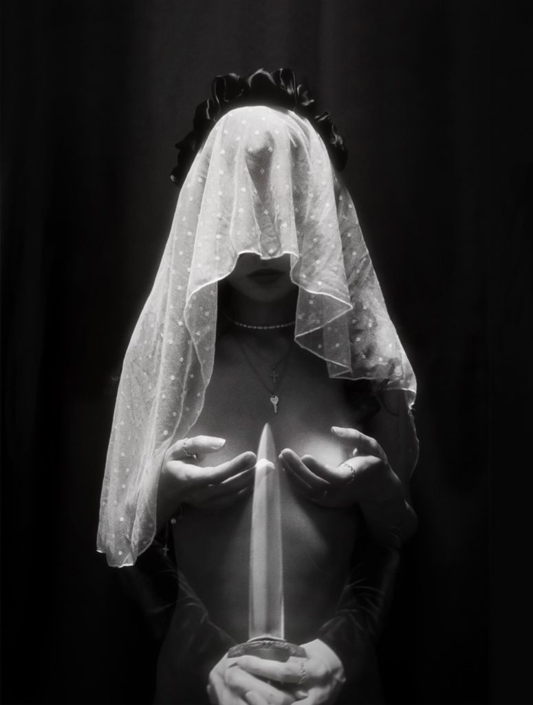 A person wearing a white veil that covers the top half of their face and a key and a crucifix around their neck holds their breasts in their hands. There is a dagger pointed upward between their breasts held by another pair of hands.